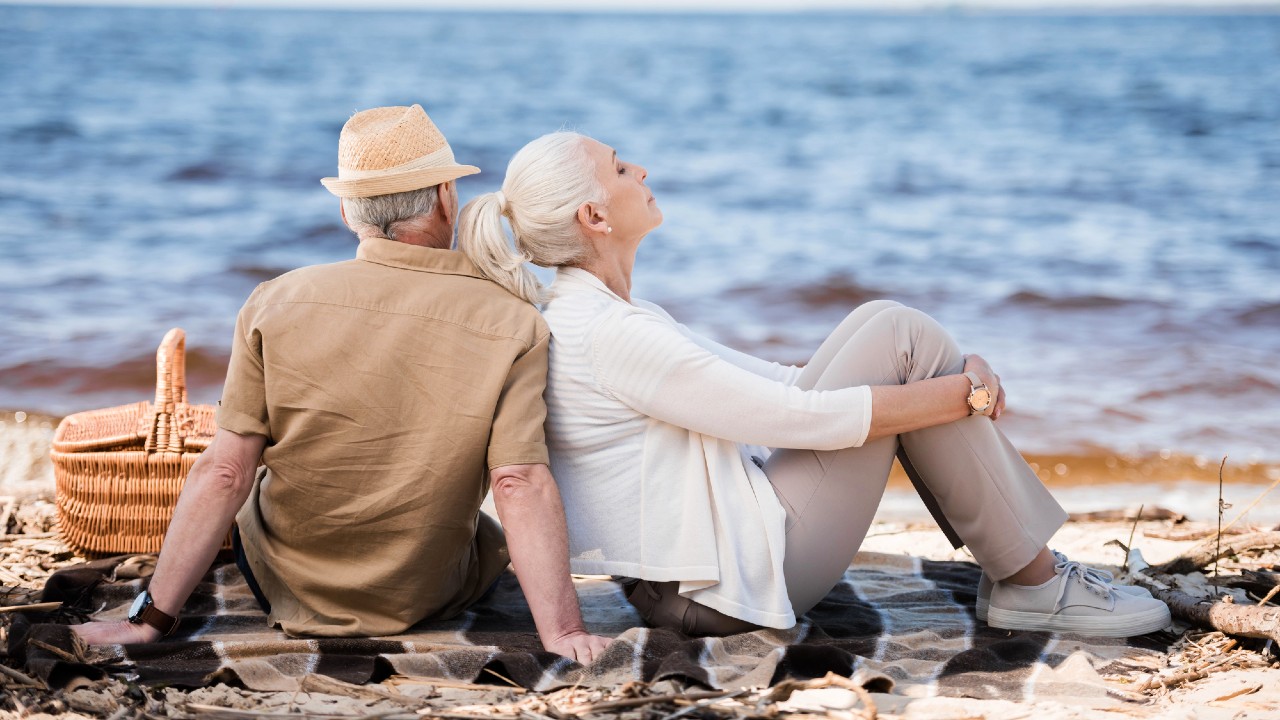 Una coppia di anziani è seduta schiena contro schiena su una coperta vicino all'acqua. L'uomo indossa un cappello e una camicia beige, e la donna ha un maglione bianco e pantaloni grigi. Un cestino da picnic è lì vicino. Sembrano rilassati e contenti sotto un cielo limpido.