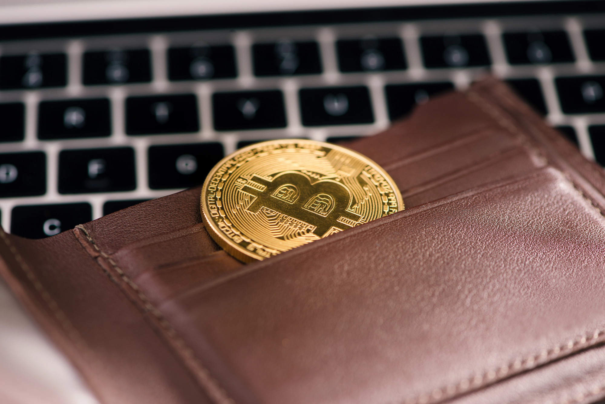 Una moneta Bitcoin d'oro sporge da un portafoglio in pelle marrone, posizionato sulla tastiera di un computer portatile.