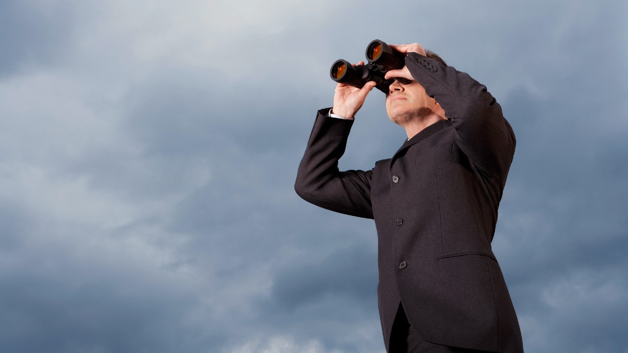 Una persona in abito scuro guarda attraverso un binocolo contro un cielo nuvoloso, apparentemente concentrata e intenta a osservare da lontano.