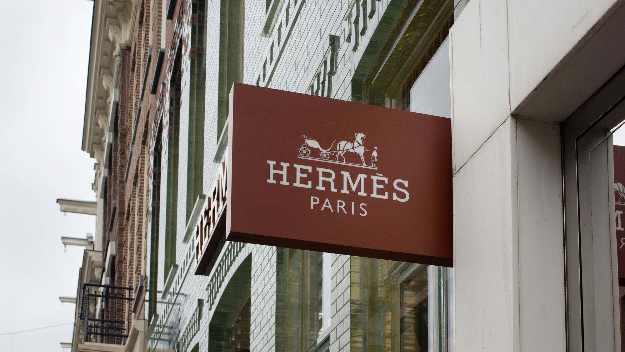 Un cartello marrone con il logo Hermes Paris, raffigurante una carrozza trainata da cavalli, è appeso all'esterno di un edificio con mattoni e pannelli bianchi. Lo sfondo mostra una vista parziale delle strutture vicine.