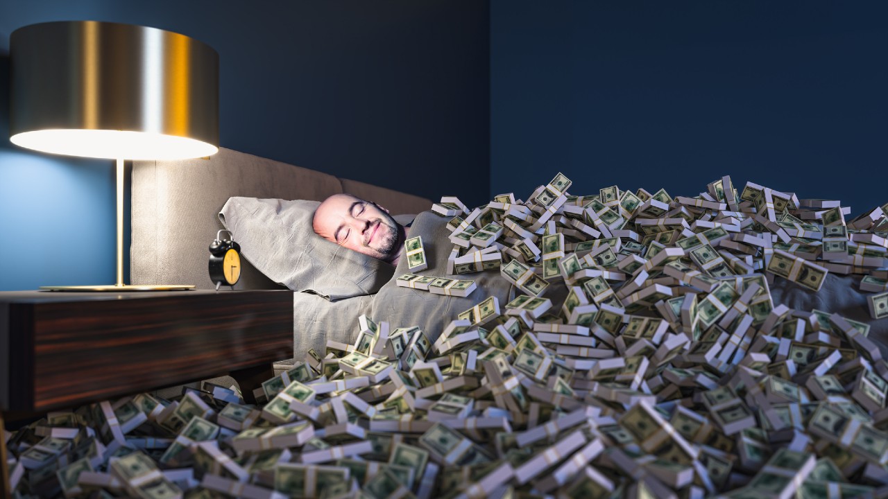 Una persona che dorme in un letto coperto da pile di banconote da un dollaro, accanto a una lampada da comodino e a un orologio. La stanza è scarsamente illuminata e ha una parete blu.