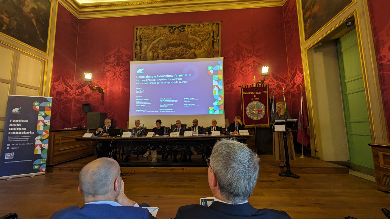 Una sala conferenze con pareti rosse e un grande schermo di proiezione che mostra una presentazione sulla cultura finanziaria. Seduti sul palco ci sono diversi relatori e un oratore è in piedi su un podio. I partecipanti sono visibili in primo piano. Striscioni e decorazioni elaborate adornano la sala.