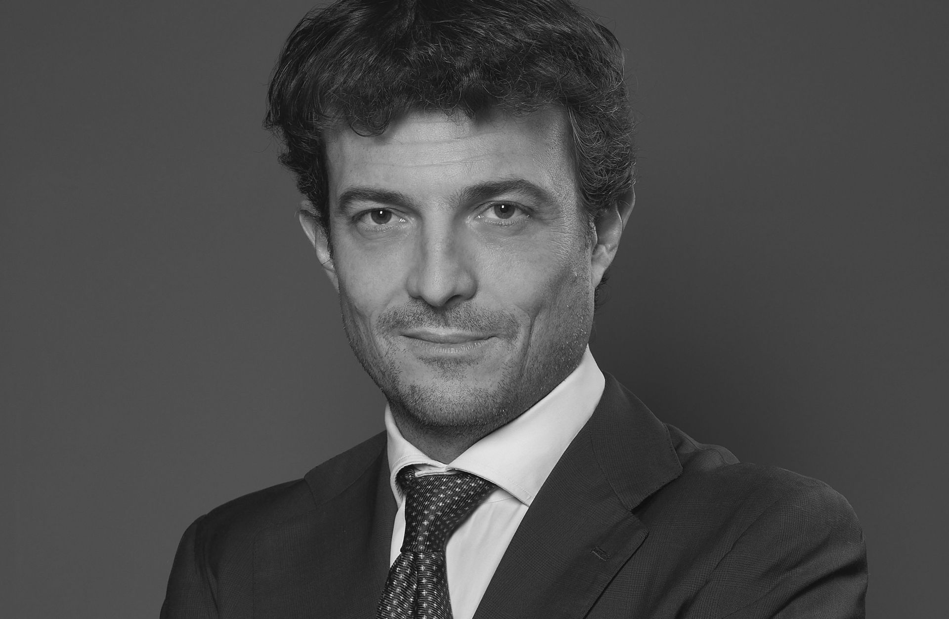 Una foto in bianco e nero di Filippo Bolaffi che parla di whisky in giacca e cravatta, con capelli ricci scuri, che guarda la telecamera. Lo sfondo è semplice e scuro, e mette in risalto l'espressione del viso e l'abbigliamento professionale.