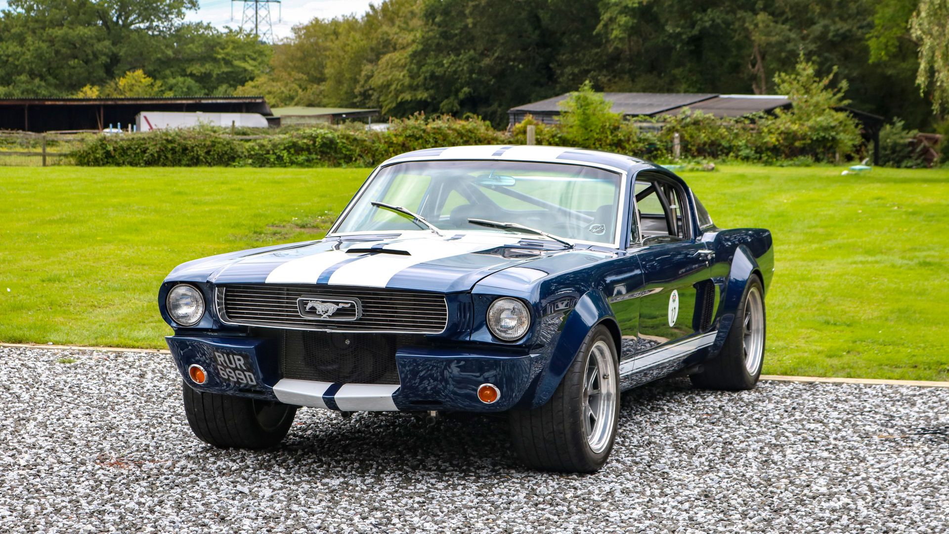 Una classica Ford Mustang blu scuro con strisce da corsa bianche è parcheggiata su una superficie ghiaiosa in un'area verde e erbosa, con alberi ed edifici sullo sfondo.