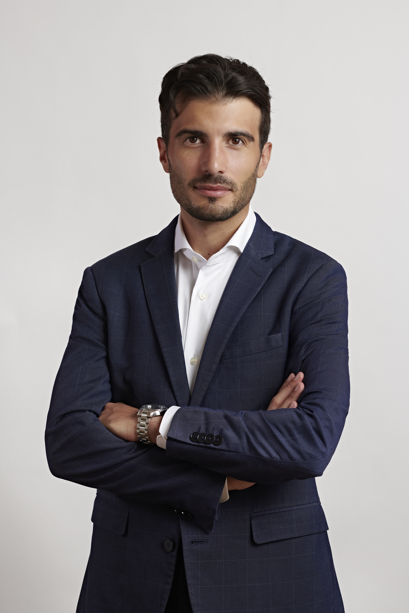 Un uomo con i capelli corti e scuri e la barba è in piedi su uno sfondo semplice. Vestito con un completo blu navy e una camicia bianca, ha le braccia incrociate, guardando direttamente la telecamera con un'espressione neutra che riflette la contemplazione del futuro delle cripto in Italia.