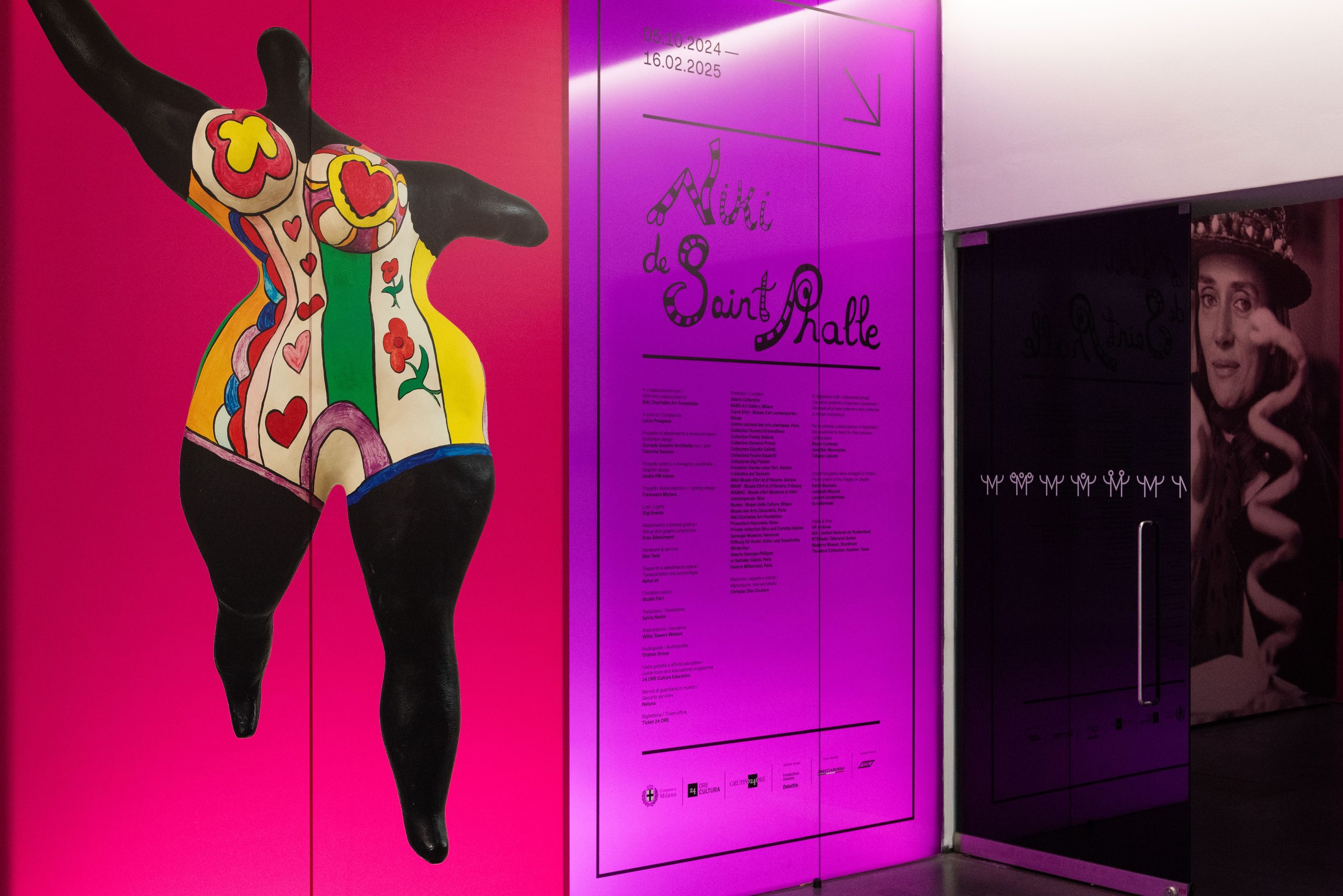 Un ingresso vivace della mostra presenta una figura femminile colorata e astratta in corsetto su uno sfondo rosa brillante. Accanto, un pannello viola condivide informazioni su "Niki de Saint Phalle", evidenziando la mostra Saint Phalle Milano. Una foto in bianco e nero è parzialmente visibile sulla destra.