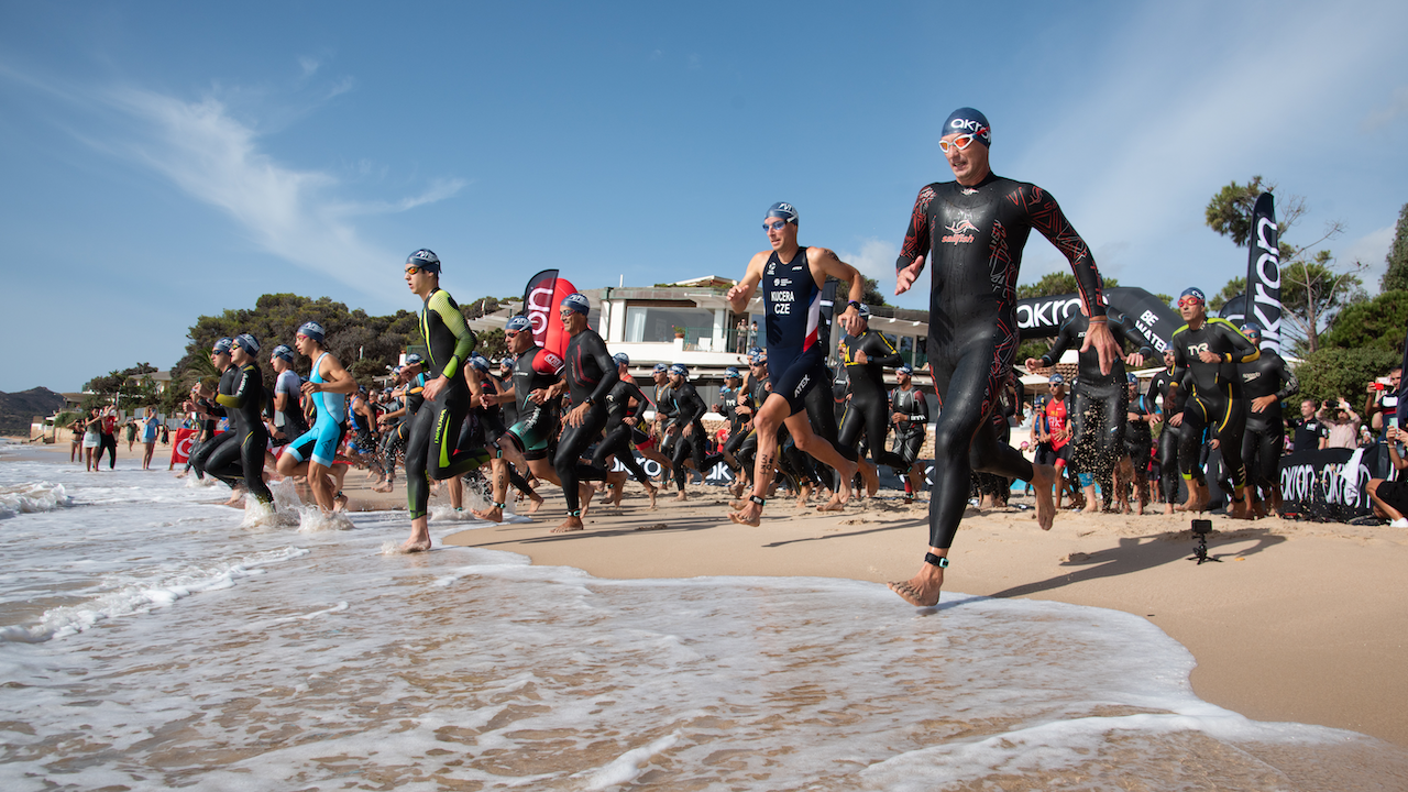 Un gruppo di triatleti in muta e cuffia da nuoto corre nell'oceano da una spiaggia sabbiosa. L'ambiente è soleggiato, con cielo terso, e ci sono spettatori sullo sfondo vicino a edifici moderni e alberi.