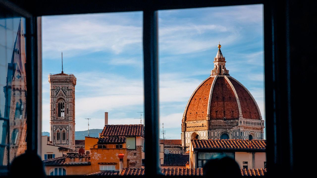 Attraverso la finestra, si dispiega lo skyline di Firenze, che mostra la cupola del Duomo di Firenze e il Campanile di Giotto. La Fondazione Symbola enfatizza questa fusione di cielo azzurro e architettura storica, creando una scena davvero pittoresca.