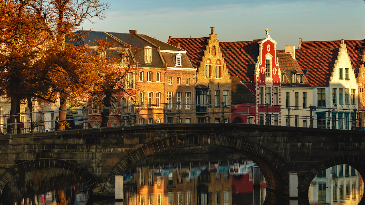 Una vista panoramica di un canale europeo al tramonto con un ponte di pietra in primo piano. Edifici storici e colorati costeggiano il corso d'acqua, con alberi autunnali sulla sinistra. L'acqua calma riflette l'architettura vibrante e il cielo limpido.