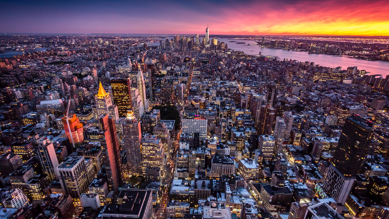 Vista aerea di un vivace paesaggio urbano al tramonto, con grattacieli densamente affollati e strade illuminate. L'orizzonte risplende di vivide tonalità arancioni e viola, mentre un fiume serpeggia attraverso la scena, riflettendo il cielo colorato.