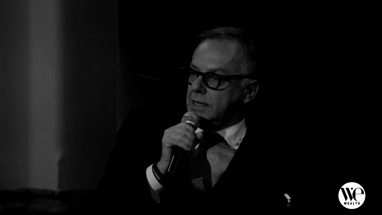 Un uomo con occhiali, giacca e cravatta parla in un microfono. L'immagine è in bianco e nero, con un logo "WE Wealth" nell'angolo in basso a destra. La scena sembra essere un evento formale o una discussione.