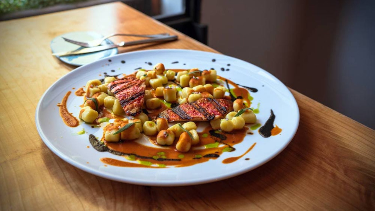 Un piatto di cibo splendidamente disposto su un tavolo di legno. Il piatto presenta fette di carne scottata circondate da gnocchi, condite con un filo di salsa e guarnite con erbe aromatiche. Una forchetta e un cucchiaio sono appoggiati accanto al piatto.