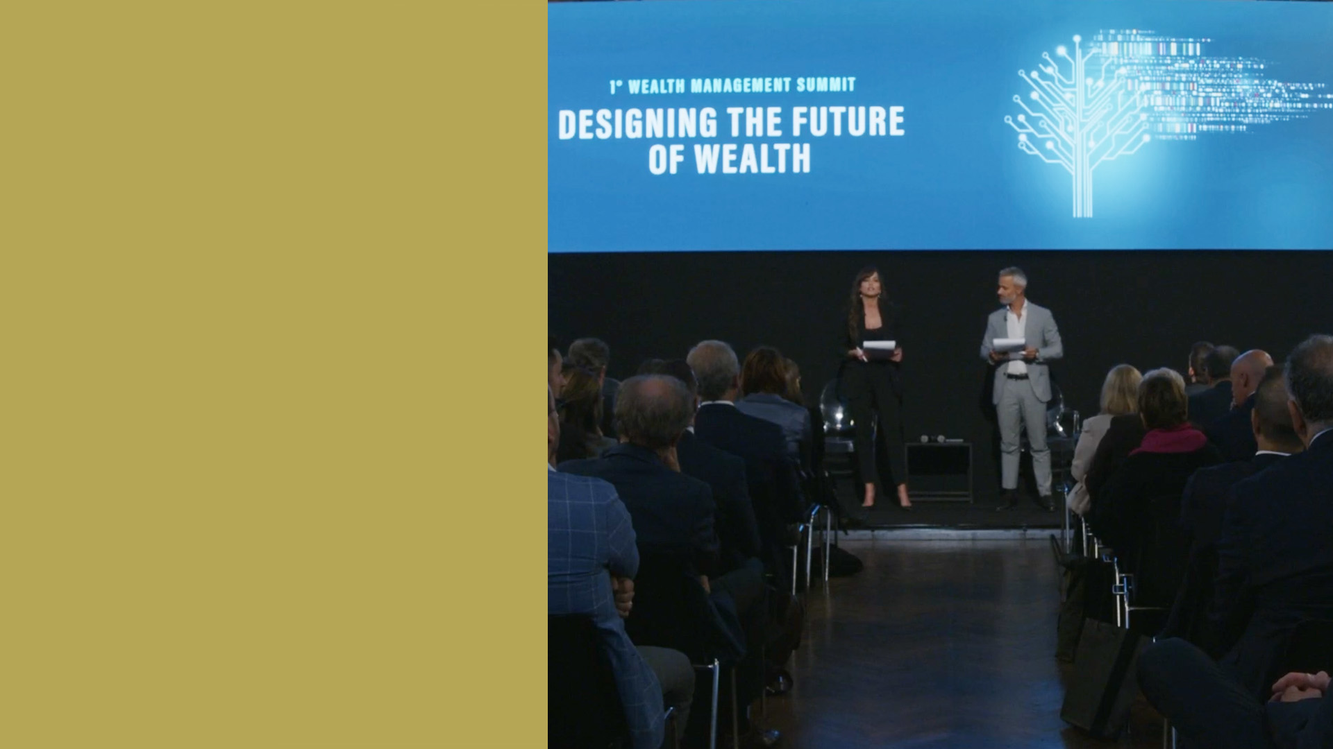 Pubblico seduto in una sala conferenze di fronte a un palco dove due relatori stanno presentando. Il grande schermo dietro di loro mostra il testo "1st Wealth Management Summit: Designing the Future of Wealth.
