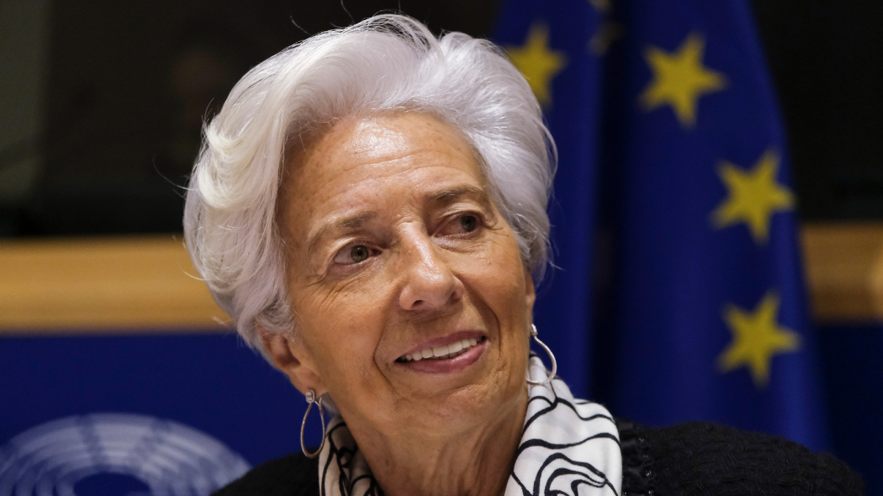 Una donna anziana con i capelli bianchi sorride leggermente, in piedi davanti alle bandiere dell'Unione Europea. Indossa una sciarpa bianca e nera e un blazer scuro.