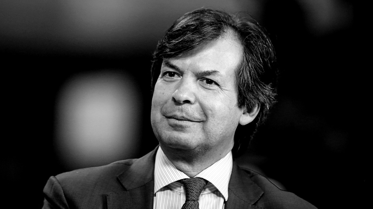 In foto, Carlo Messina, ceo di Intesa Sanpaolo, in giacca e cravatta, sorride leggermente. L'immagine è in bianco e nero, con sfondo sfocato.