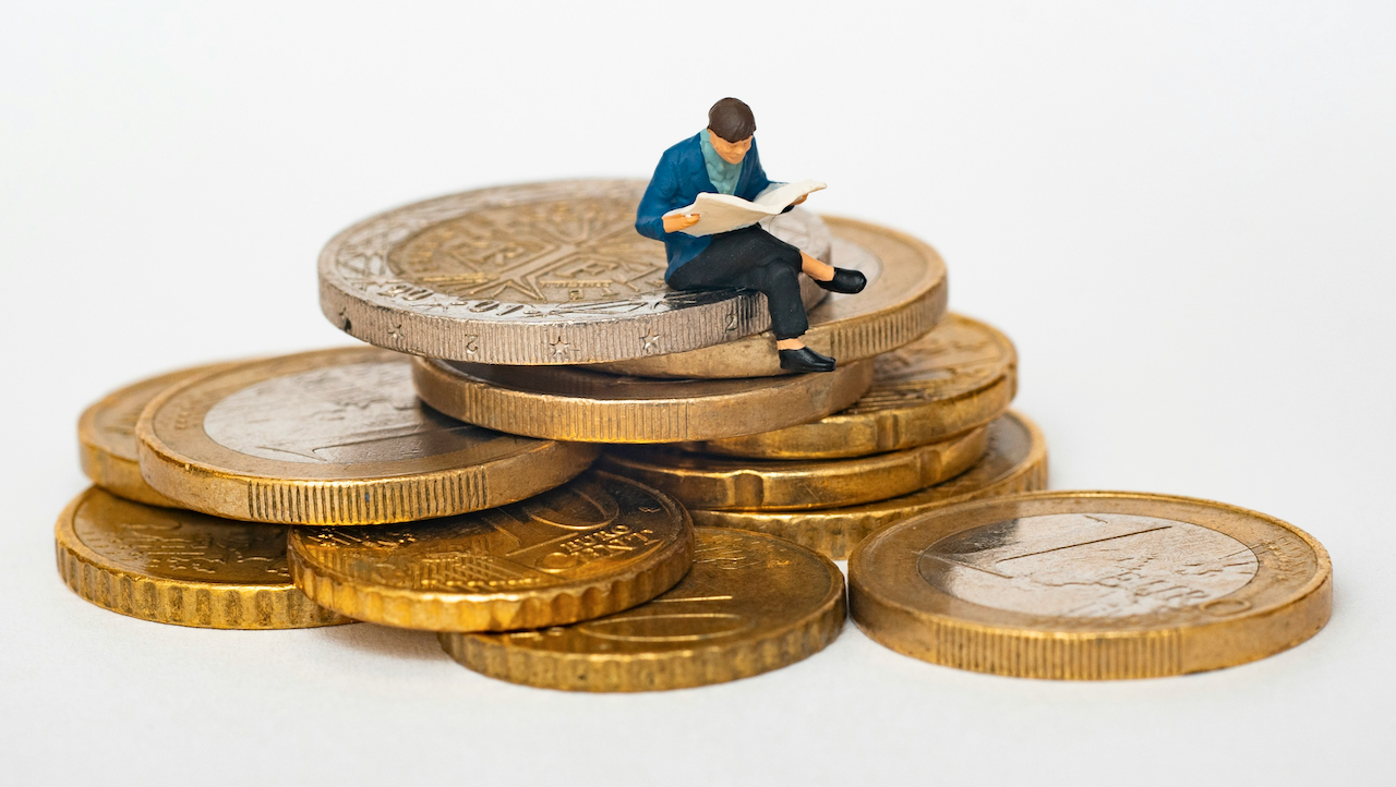 Una piccola statuetta raffigurante una persona in blu è seduta su una pila di monete e legge un giornale. La scena presenta grandi monete d'oro e d'argento disposte in una pila, evidenziando i temi della finanza e degli investimenti.