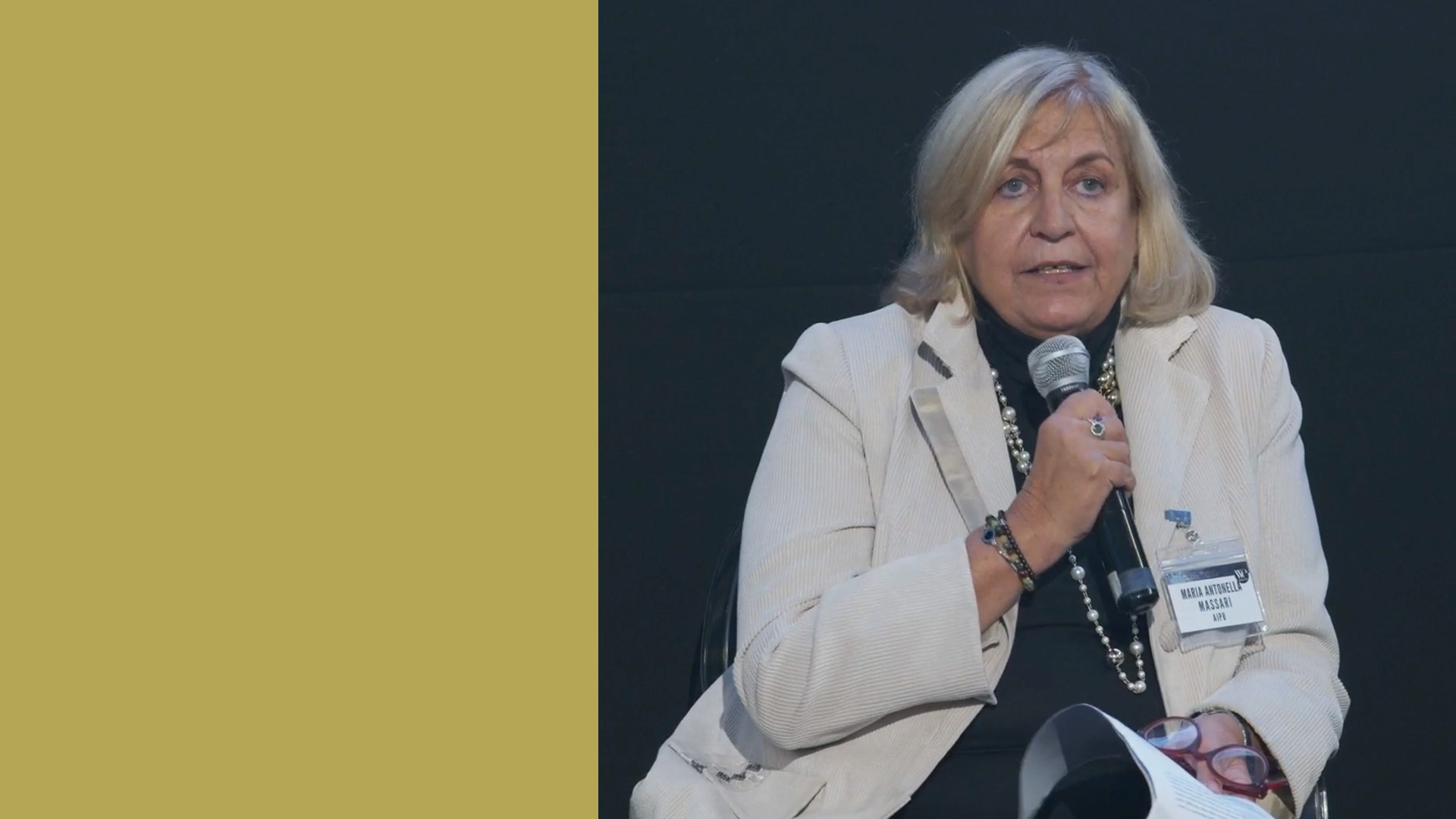 Antonella Massari, segretario generale di AIPB, indossa un blazer beige chiaro e tiene in mano un microfono, è seduta su uno sfondo scuro. Sembra che stia parlando. C'è uno spazio vuoto verde oliva alla sua sinistra.