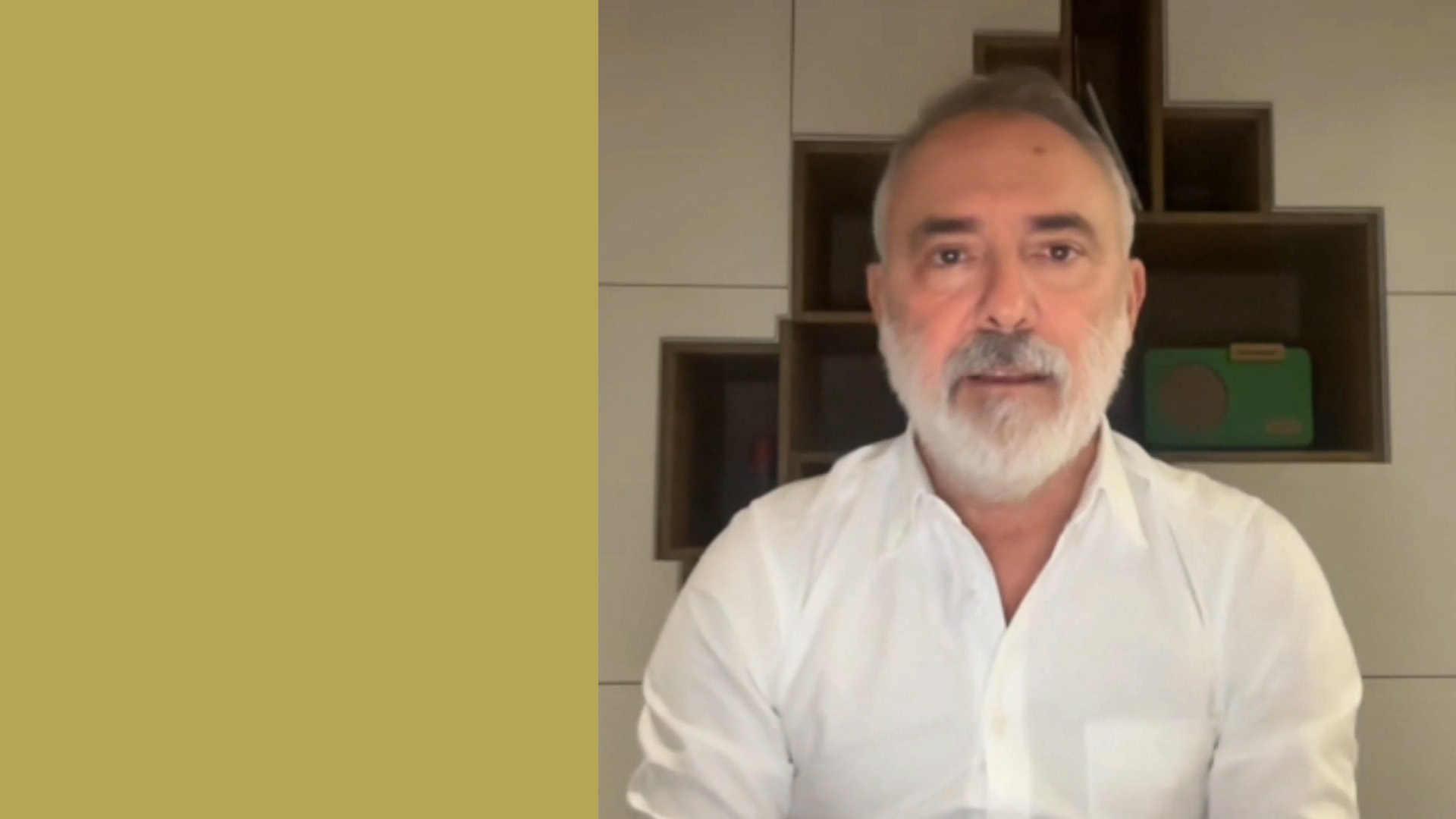 Un uomo con barba e baffi indossa una camicia bianca, seduto davanti a una mensola geometrica. Lo sfondo è diviso, con un colore giallo senape sul lato sinistro e un interno moderno sulla destra.