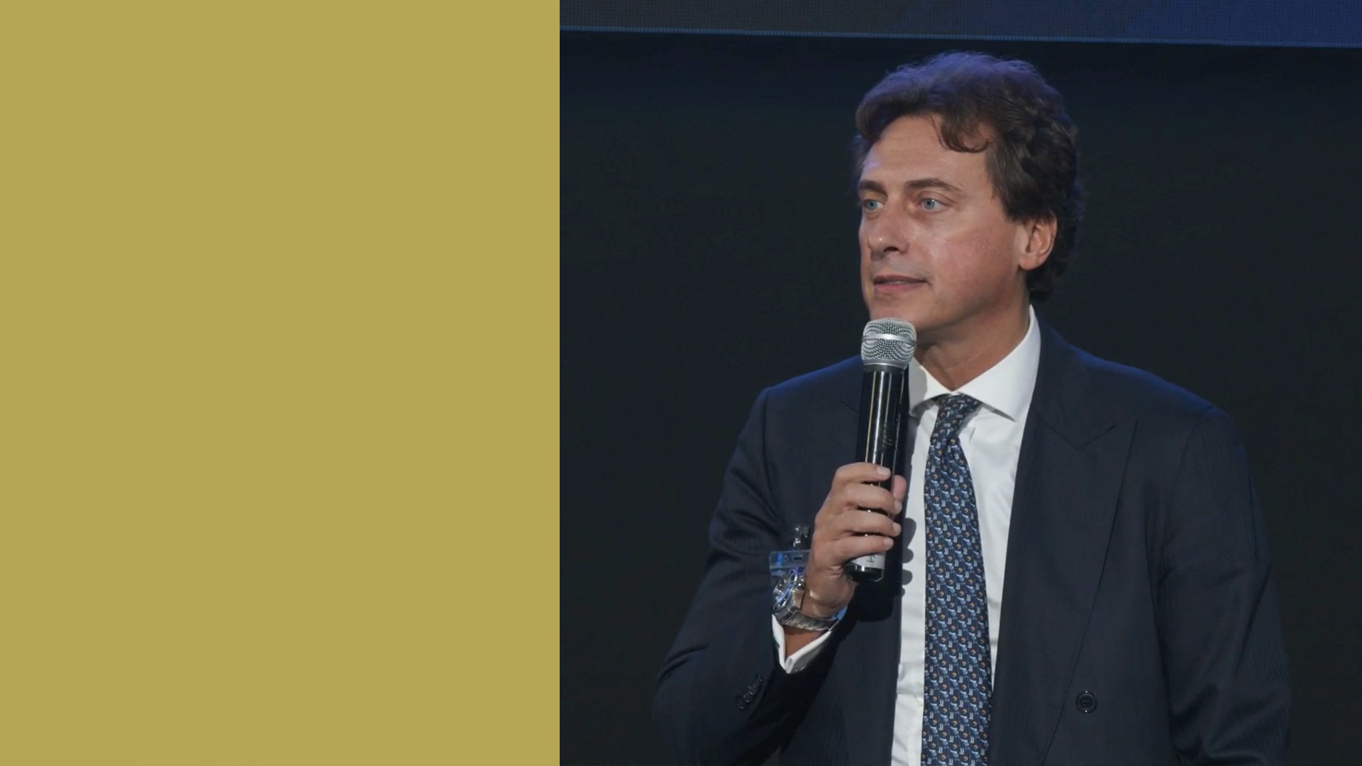 Un uomo in giacca e cravatta, che rappresenta l'era bancaria, tiene un microfono mentre parla sul palco contro uno sfondo nero. Sulla sinistra, c'è una striscia verticale color senape, che crea un contrasto sorprendente.
