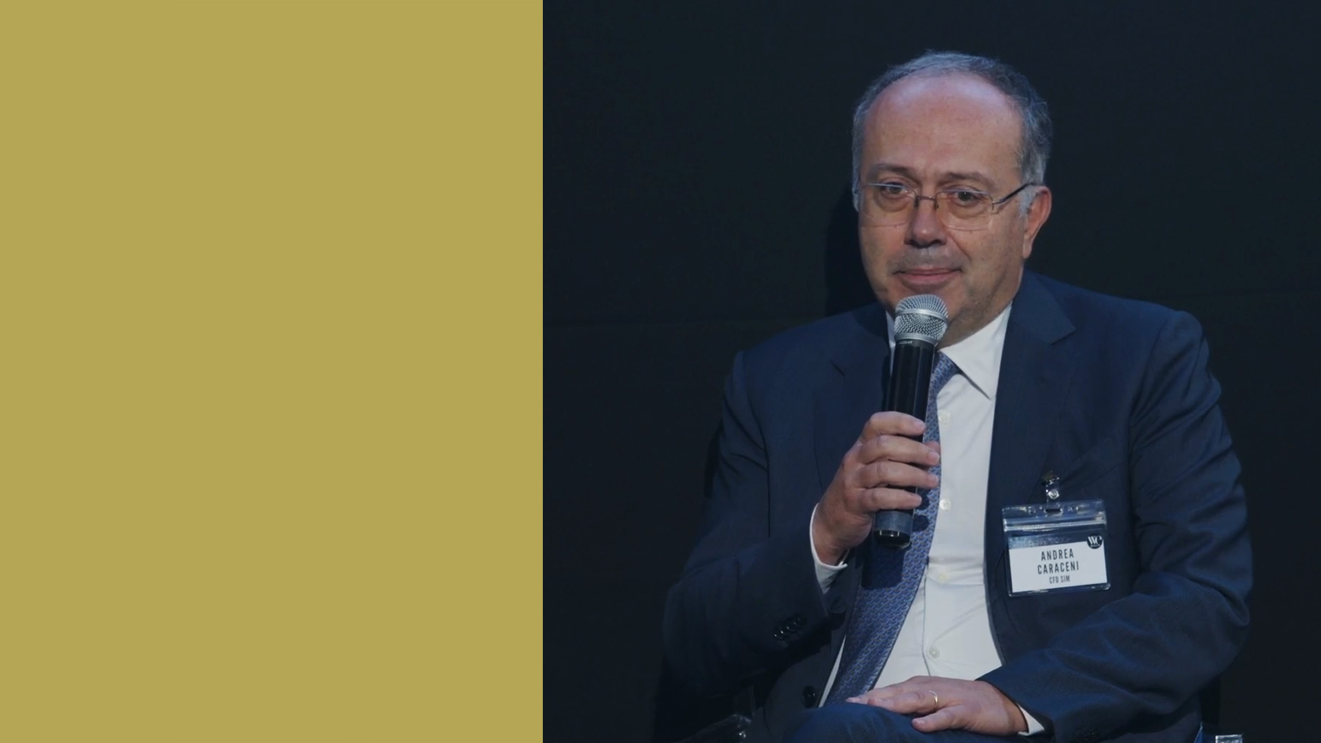 Andrea Caraceni parla al microfono seduto, incarnando la professionalità raffinata tipica di un dirigente di Family Office. Con occhiali e un cartellino identificativo, siede su uno sfondo prevalentemente nero con una vistosa sezione giallo senape sul lato sinistro.