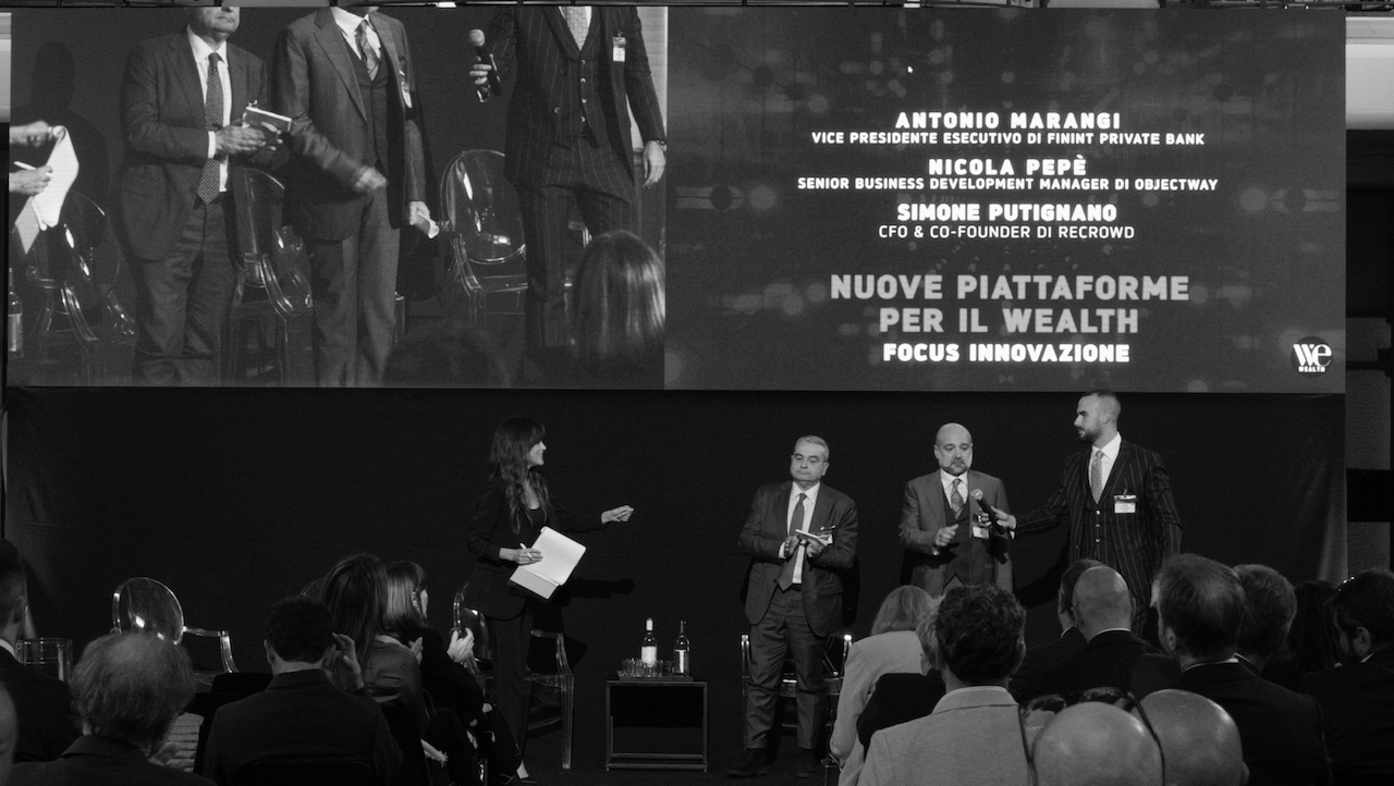 Un'immagine in bianco e nero di una conferenza aziendale. Quattro relatori sono sul palco con i microfoni in mano, con uno schermo dietro di loro che mostra i loro nomi e ruoli. Il pubblico è seduto, di fronte al palco.