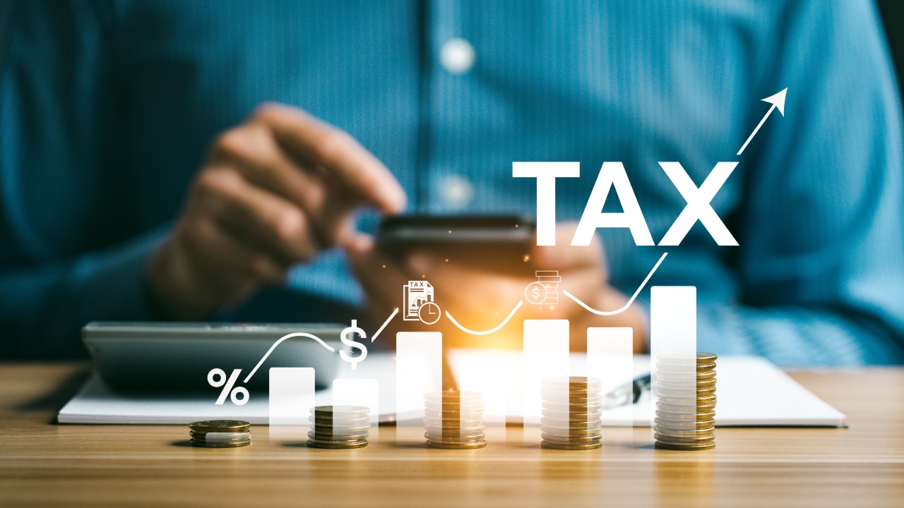 Una persona con una camicia blu usa uno smartphone, seduta a una scrivania con monete impilate che rappresentano la successione e la crescita finanziaria. Le icone bianche mostrano segni di percentuale e simboli del dollaro, mentre "TAX" appare in grassetto accanto a una freccia rivolta verso l'alto, illustrando concetti economici chiave.