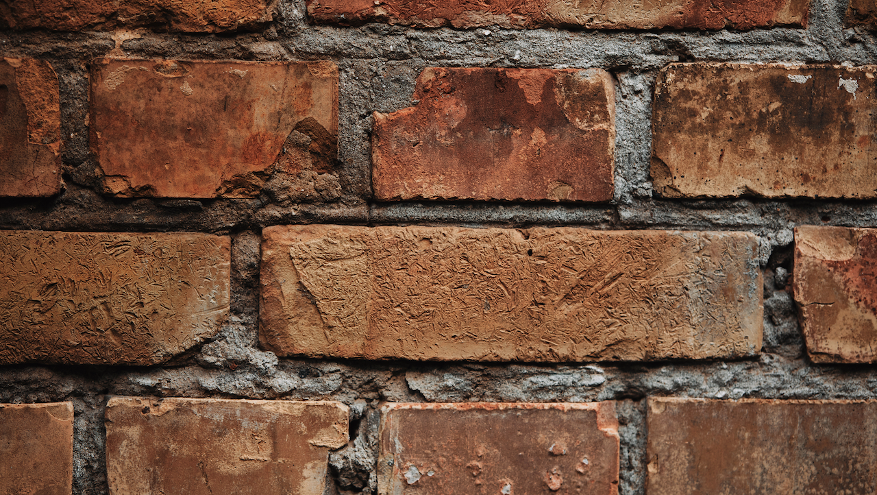 Primo piano di un muro di mattoni rosso-marrone con linee di malta visibili. Alcuni mattoni sono scheggiati e hanno una superficie ruvida, che mostra segni di usura e di età. L'illuminazione mette in risalto le varie tonalità e texture dei mattoni