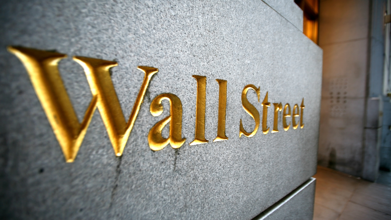 Primo piano di un muro di pietra con la scritta "Wall Street" in rilievo in lettere dorate. Il testo è angolato, creando un senso di profondità e prospettiva. L'area circostante appare urbana e ben illuminata.