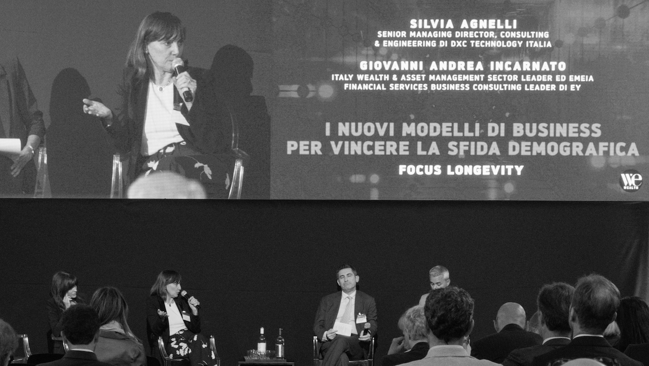 Una foto in bianco e nero che mostra una tavola rotonda. Quattro persone sono sedute su un palco in un contesto di conferenza. Un grande schermo sopra mostra nomi e titoli. L'evento si concentra su modelli di business e sfide demografiche.