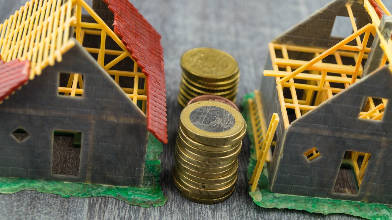 Due piccole case modello con tetti incompleti sono appoggiate su una superficie grigia. Una pila di monete in euro, che ricordano potenziali strategie finanziarie come la prossima manovra 2025, si trova tra di loro, a simboleggiare l'investimento o il costo in costruzione o immobiliare.