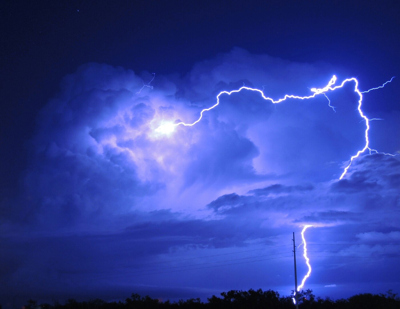 Un vivido fulmine illumina un cielo notturno scuro e nuvoloso. Il lampo luminoso contrasta con il blu intenso, evidenziando le drammatiche nubi temporalesche. In primo piano sono visibili le sagome di alberi lontani e una linea elettrica.
