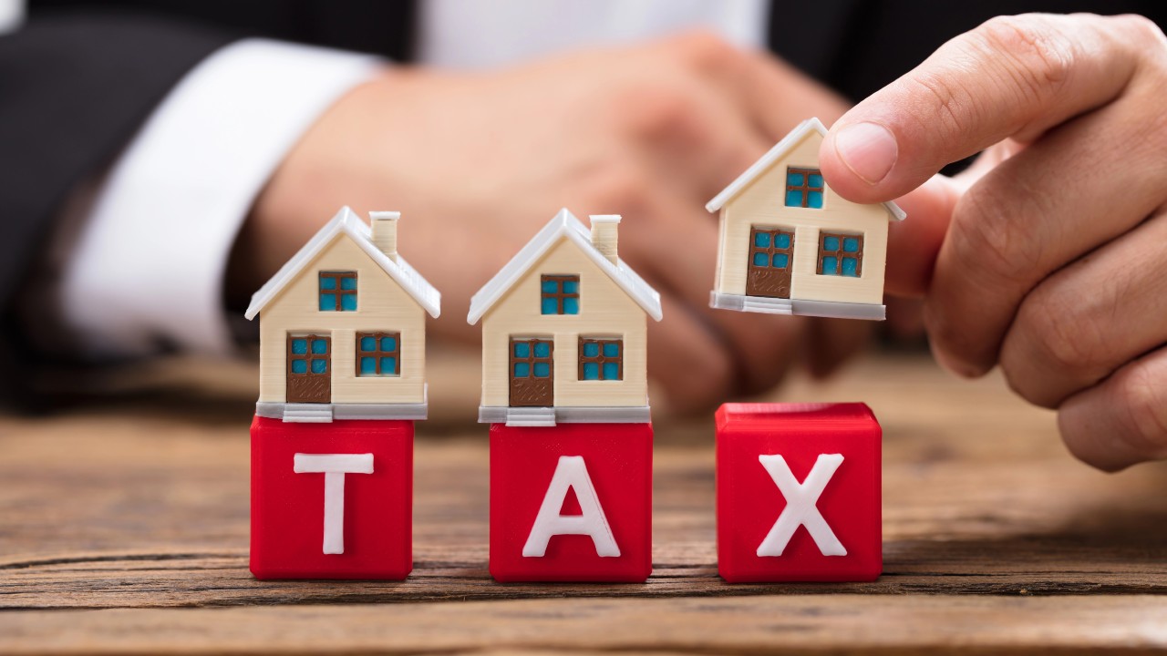 Una fila di case in miniatura è posta in cima a blocchi rossi con la scritta "TAX" su un tavolo di legno. La mano di una persona tiene una delle case, che simboleggia le tasse sulla proprietà o gli investimenti immobiliari, evidenziando il concetto di residenza fiscale per determinare dove sono applicabili le tasse.