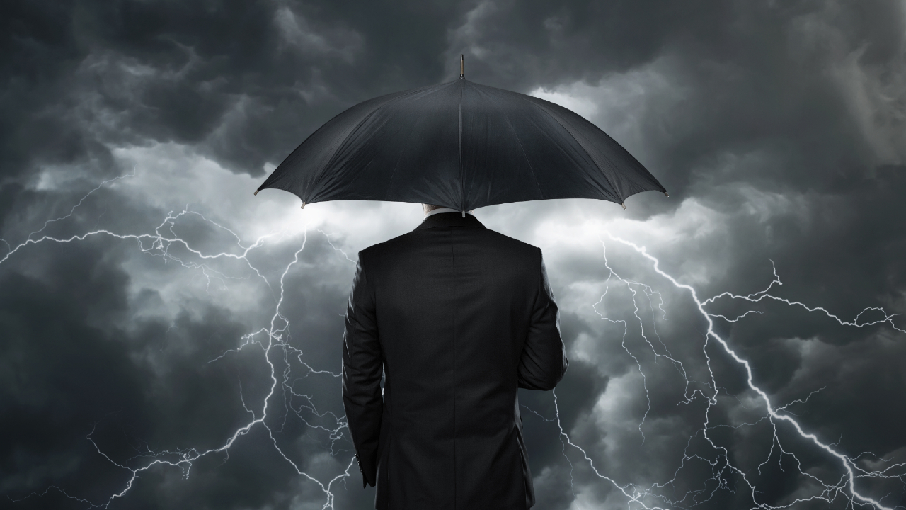 Una persona in abito è in piedi, con le spalle rivolte alla telecamera, e tiene in mano un ombrello nero, mentre lo sguardo si staglia contro un cielo drammatico, pieno di nuvole scure e fulmini luminosi, abbracciando il risiko che danza nella tempesta.