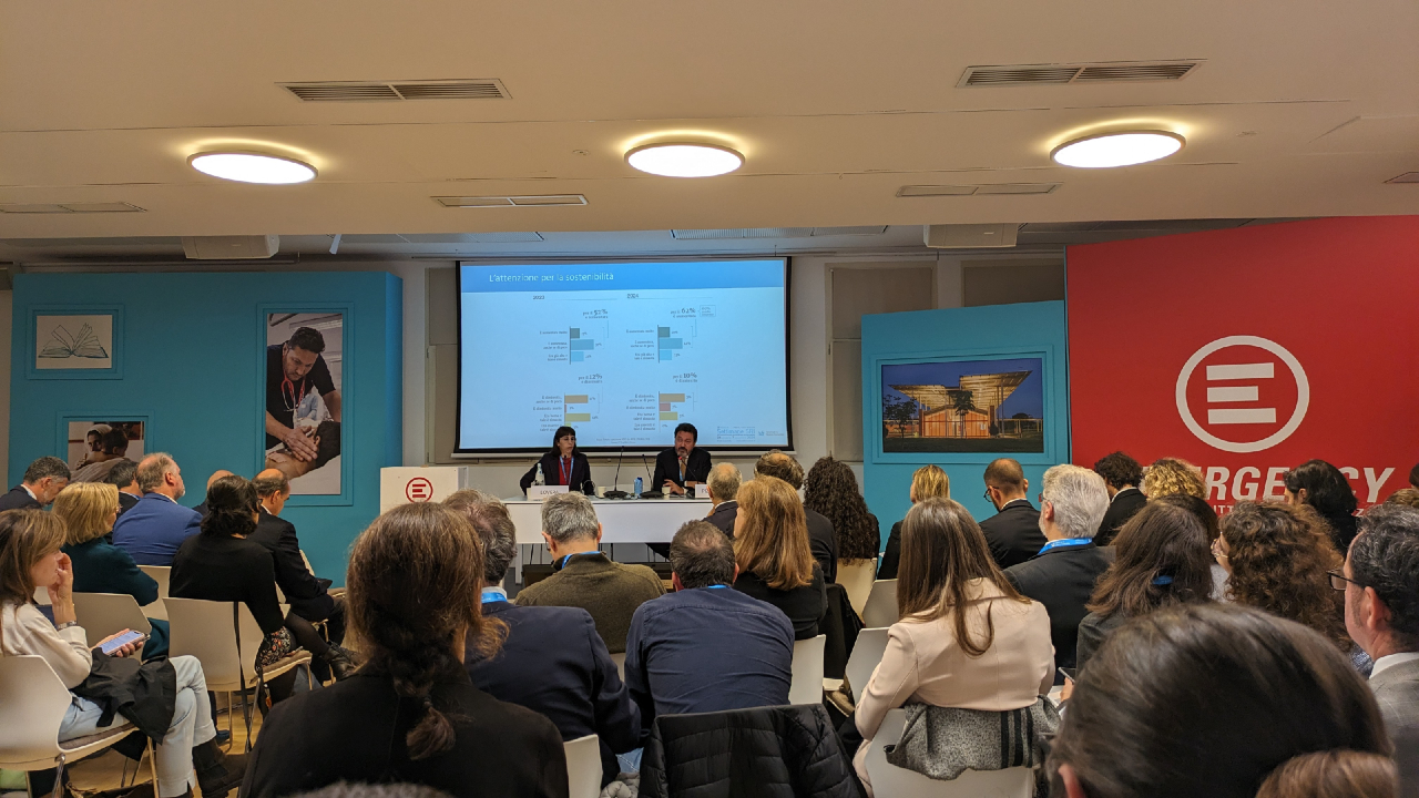 Una sala seminari piena di attesa ospita un vasto pubblico seduto e concentrato su due relatori che discutono di "sostenibilità" al tavolo di fronte. Una presentazione lampeggia sullo schermo dietro di loro, mentre le pareti espongono poster e un sorprendente pannello rosso decorato con un logo bianco.