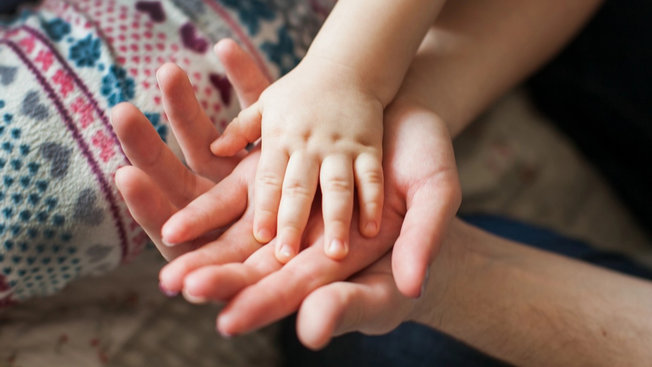 La mano di un bambino poggia delicatamente sul palmo della mano di un adulto, a simboleggiare una successione di generazioni. Un tessuto lavorato a maglia colorato è visibile sullo sfondo, aggiungendo calore al tenero momento. L'idea ricorda il passaggio generazionale, legato al tema della successione e della donazione