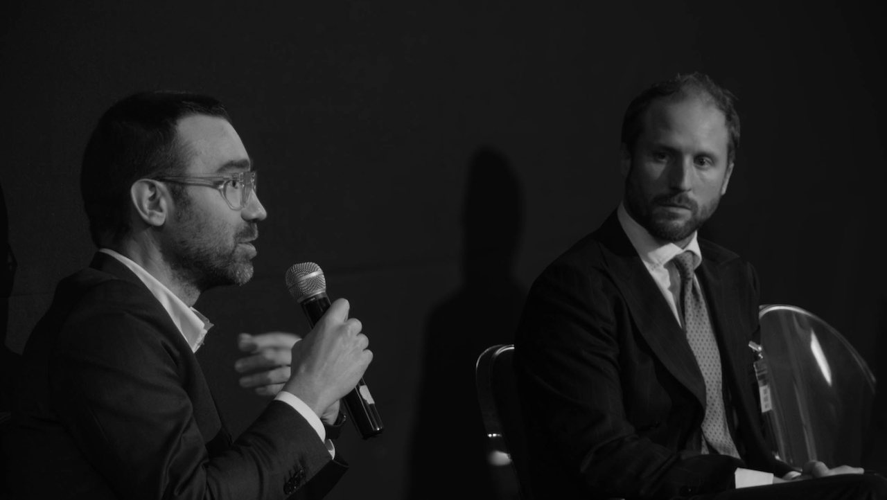 Due uomini in giacca e cravatta, Luca Giacobbe e Tommaso Migliore, sono impegnati in una conversazione, discutendo delle ultime novità in fatto di tecnologia. Uno parla al microfono mentre l'altro ascolta attentamente. L'ambiente è formale, con uno sfondo scuro e un riflettore che mette in risalto gli oratori.