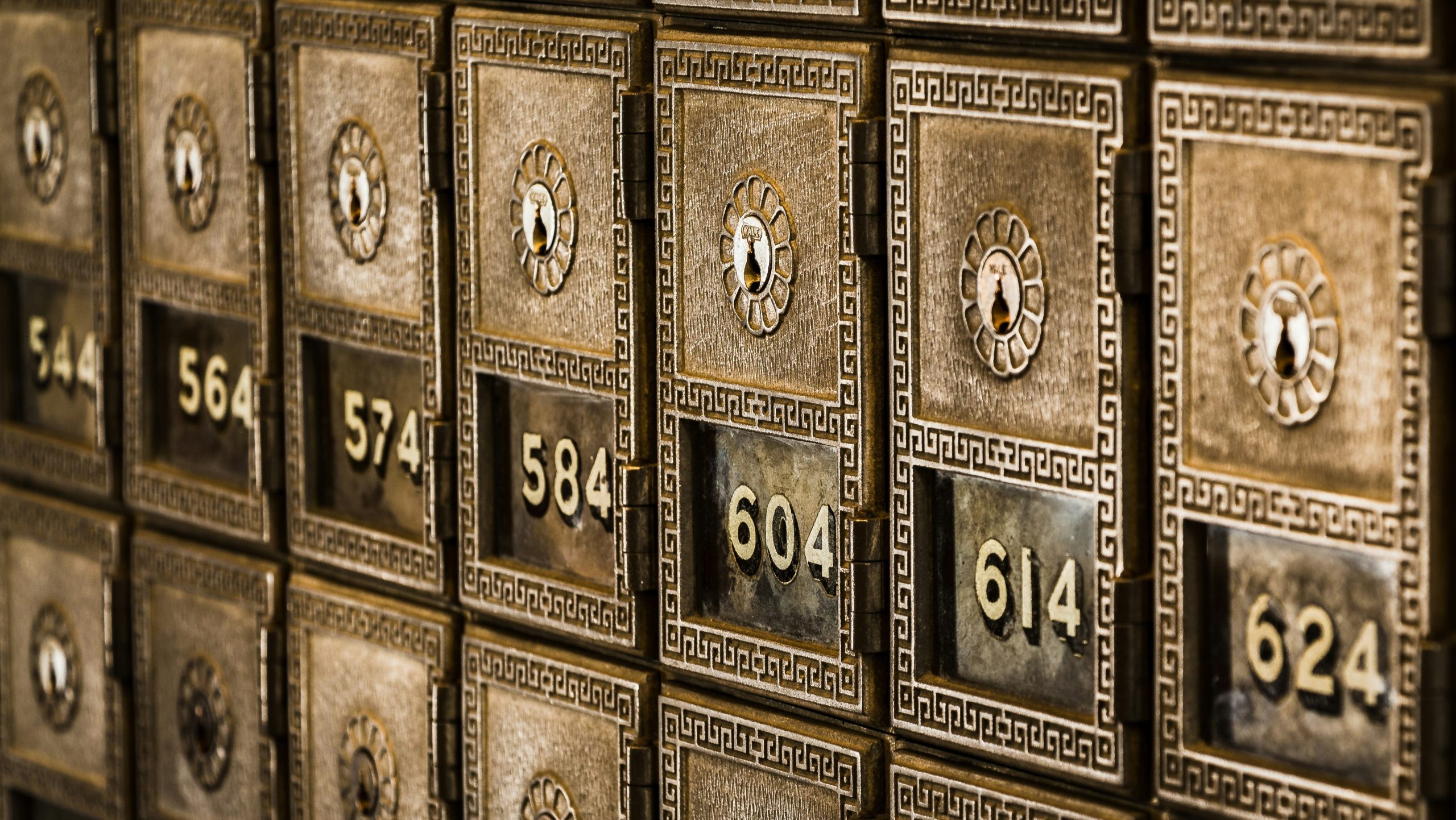 Primo piano di cassette postali d'ottone d'epoca con lucchetti a combinazione e motivi ornamentali. Ogni cassetta è numerata, con numeri visibili tra cui 564, 574, 584, 604 e 614. Le cassette hanno buchi della serratura decorati al centro.