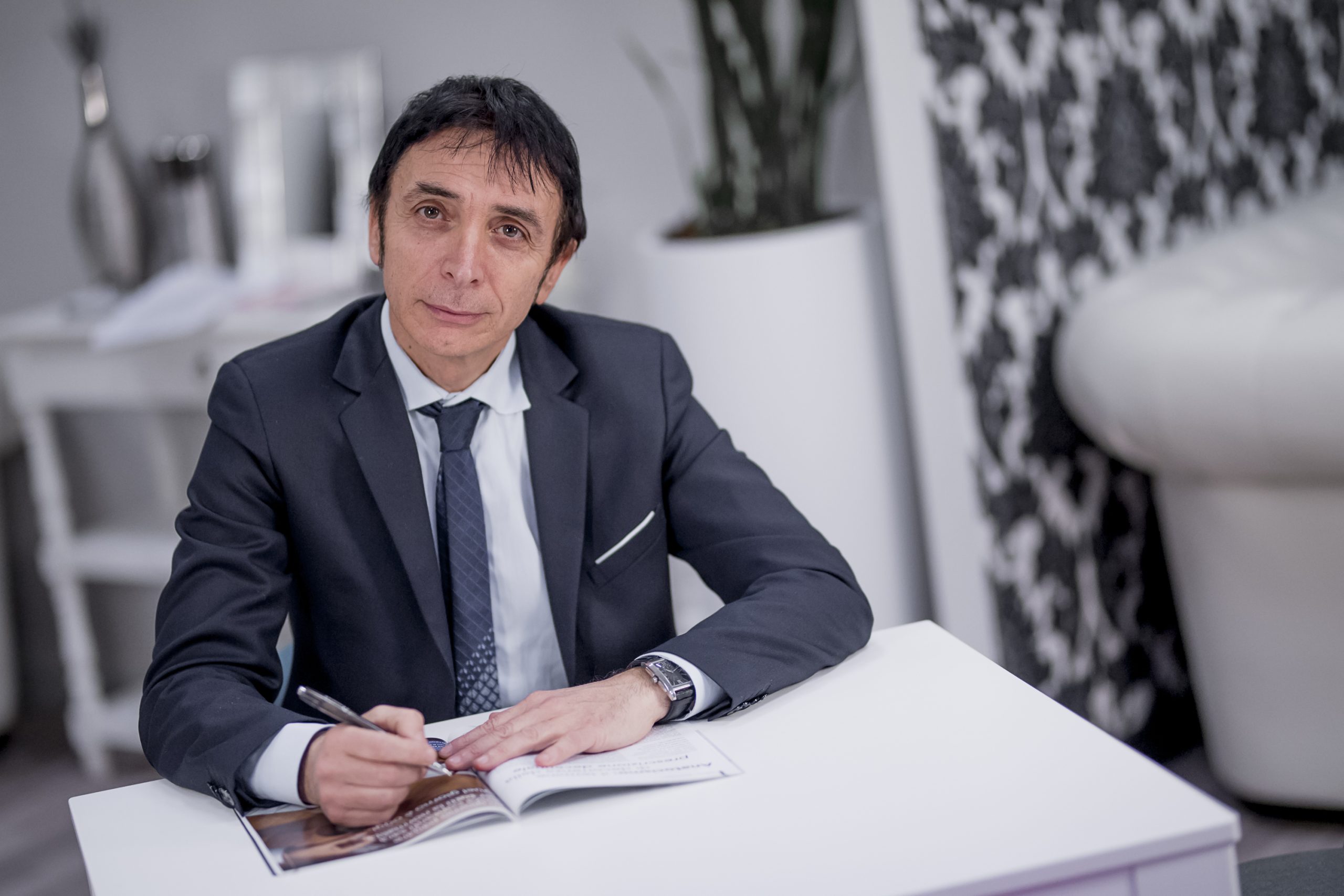 Un uomo in giacca e cravatta è seduto a una scrivania bianca, con una penna in mano e un quaderno su cui scrivere. Sullo sfondo ci sono vasi decorativi, una pianta e una parete decorata.