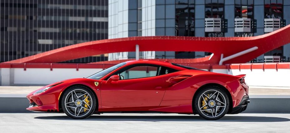 2020 Ferrari F8 Tributo. Un'auto sportiva rossa è parcheggiata su un tetto con un grattacielo moderno ed elegante sullo sfondo. L'auto presenta pinze dei freni gialle e cerchi neri distintivi. Un elemento architettonico rosso si curva sopra l'auto, aggiungendo all'ambiente urbano.