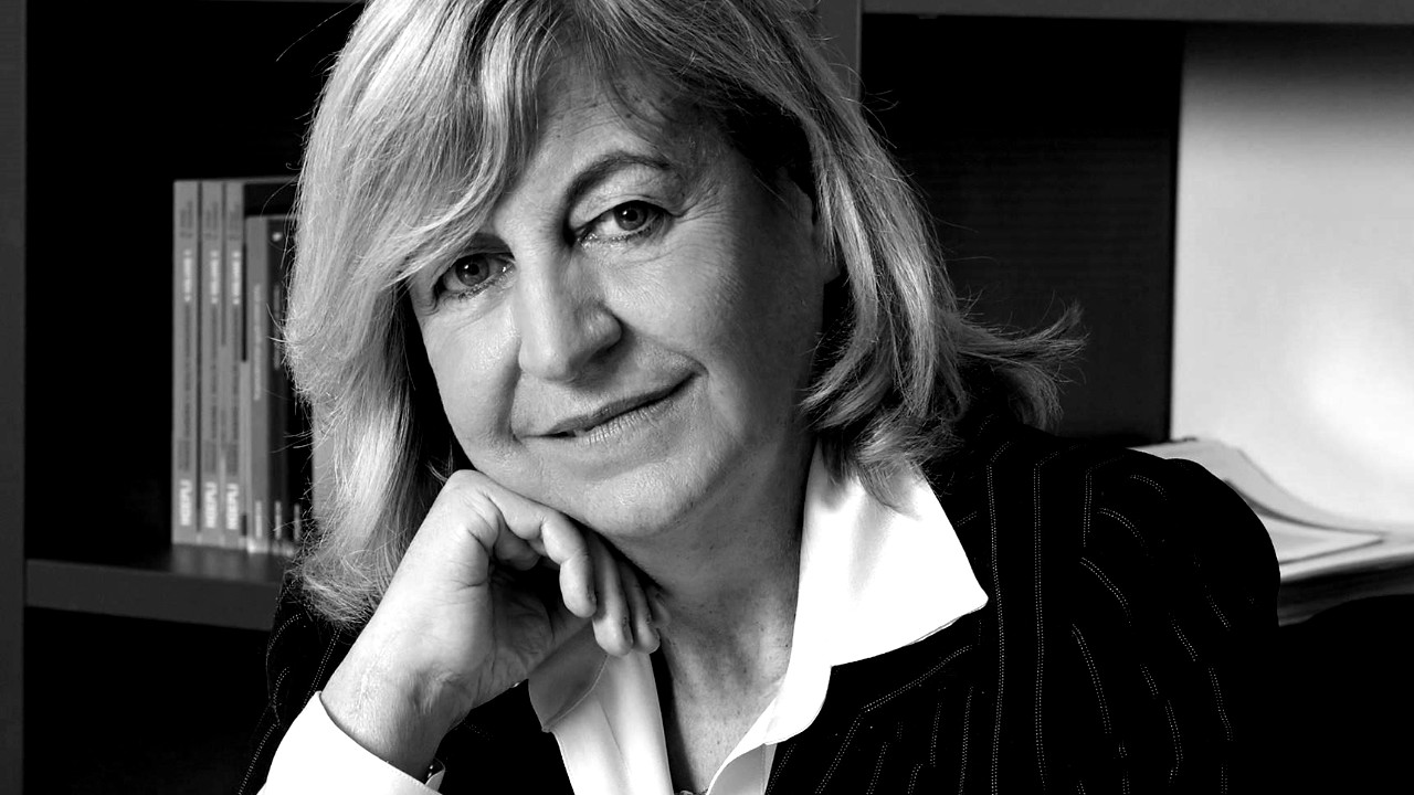 Ritratto in scala di grigi di una persona con i capelli lunghi fino alle spalle, che indossa una giacca e una camicia bianca. Appoggia il mento su una mano e guarda la telecamera, con dei libri visibili sullo sfondo.