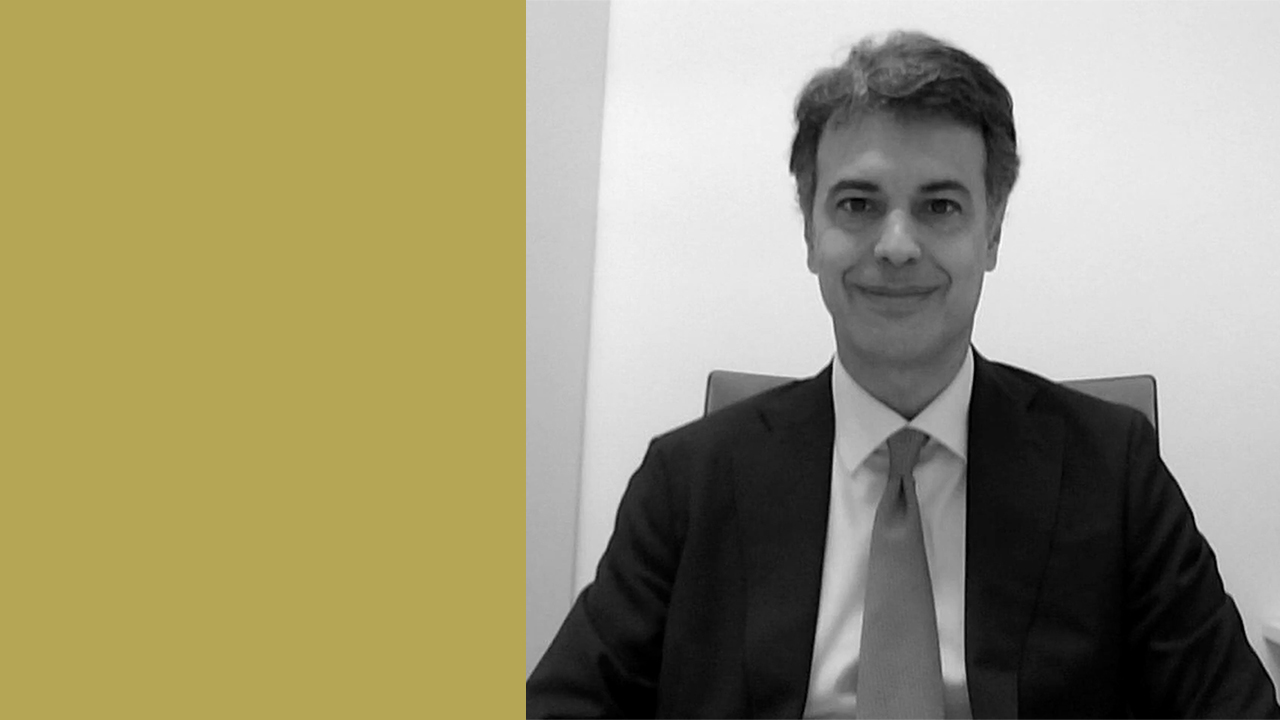 Un'immagine in scala di grigi di un uomo in giacca e cravatta, ovvero l'avvocato Francesco Capitta dello studio Rossi Facchini Michelutti, seduto su una sedia da ufficio, che sorride alla telecamera. C'è un grande rettangolo dorato verticale sul lato sinistro dell'immagine.