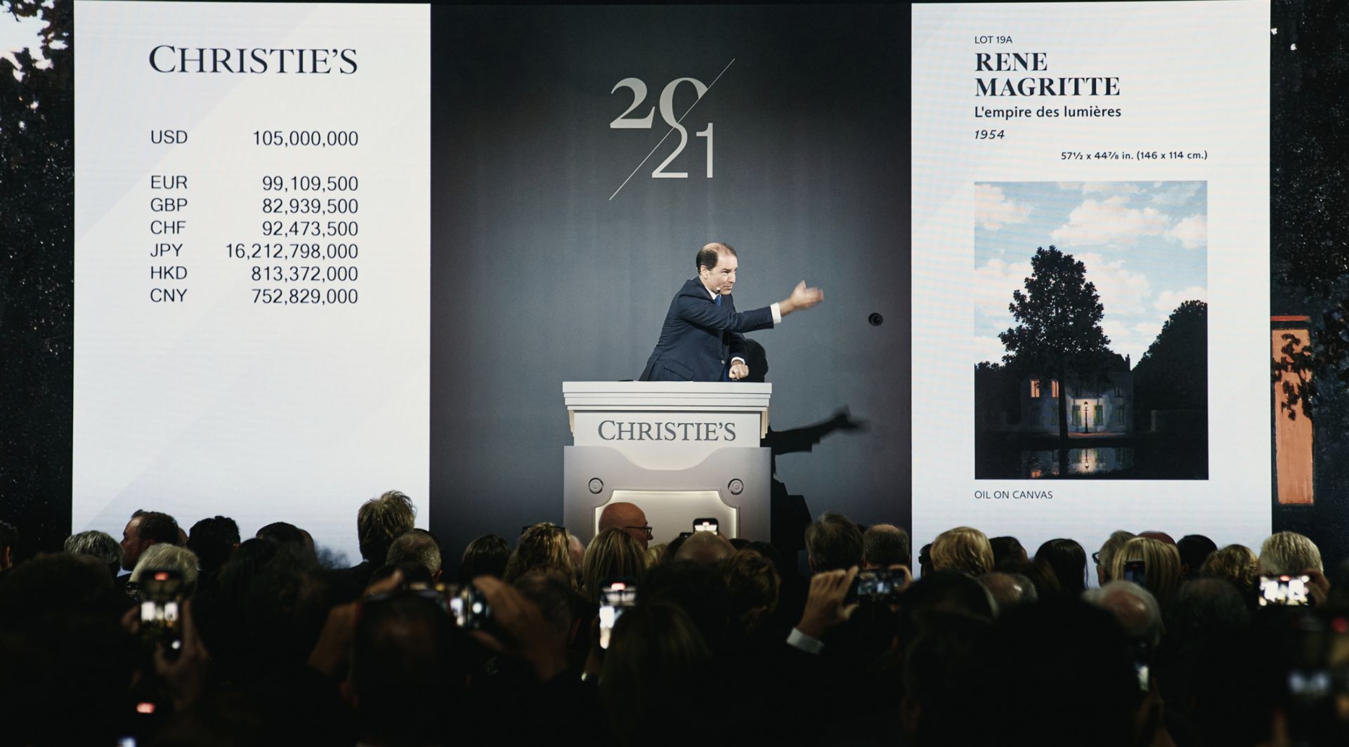 Un banditore di Christie's è in piena azione, mentre supervisiona un'asta movimentata. Uno schermo mostra il dipinto di René Magritte "L'empire des lumières" con un prezzo di partenza di 105.000.000 di USD. Il pubblico tiene in mano i telefoni, catturando il momento.