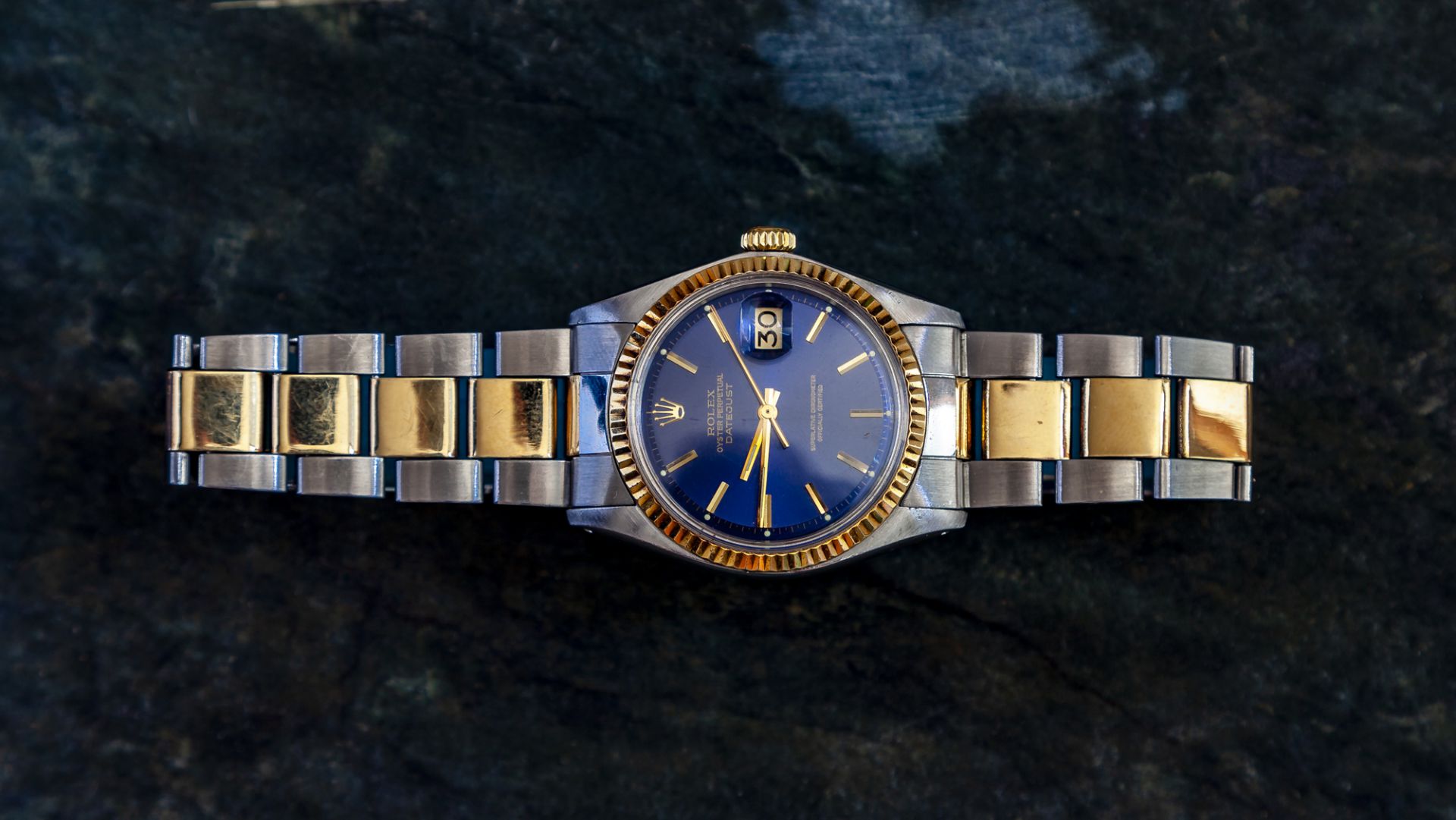Come va il mercato degli orologi? Un orologio di lusso con quadrante blu e accenti dorati è esposto su uno sfondo scuro testurizzato. Presenta un cinturino in metallo con maglie dorate alternate e una finestrella per la data alle ore tre.