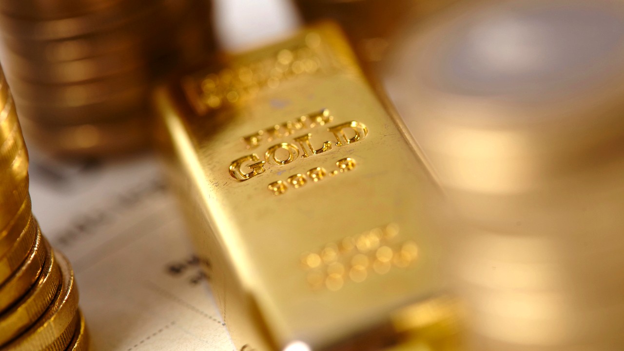 Primo piano di una barra d'oro con inciso "999.9" circondata da pile di monete d'oro. Lo sfondo presenta testo sfocato e documenti finanziari, suggerendo un'attenzione rivolta all'investimento o alla finanza.