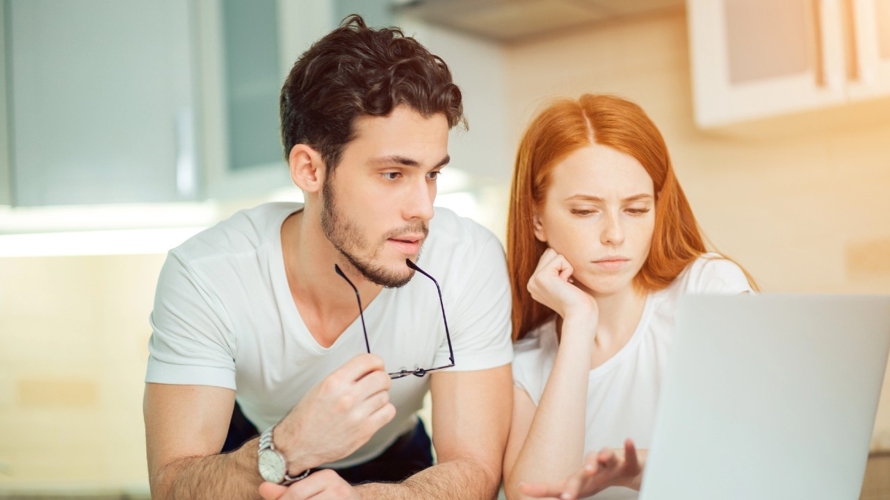 Un uomo e una donna sono concentrati sullo schermo di un computer portatile in una cucina. L'uomo tiene in mano degli occhiali e indossa una maglietta bianca, mentre la donna, anche lei con una maglietta bianca, appoggia il mento sulla mano. Entrambi hanno un'espressione concentrata.