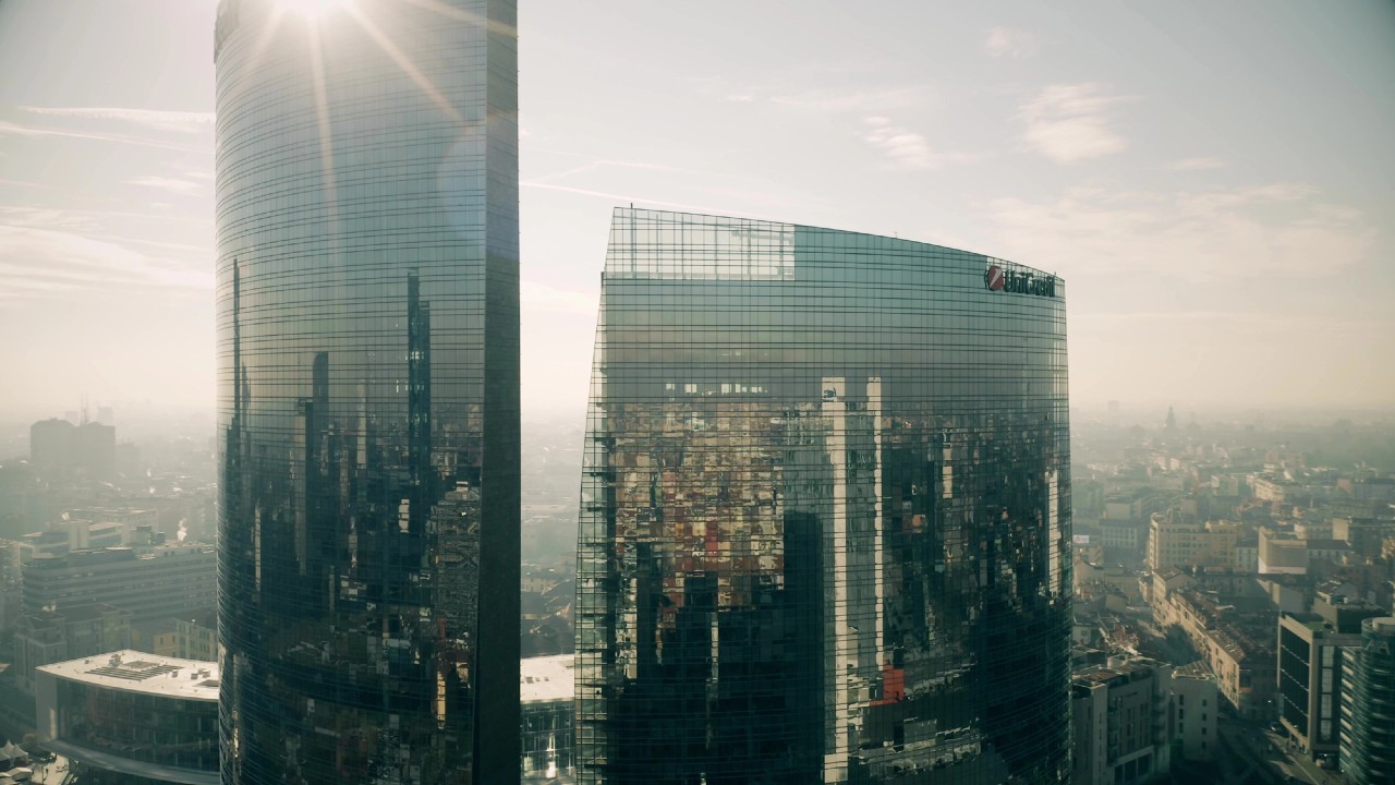 Alti grattacieli moderni con facciate in vetro riflettente svettano su un paesaggio urbano. Gli edifici riflettono l'ambiente urbano e la luce del sole, creando un motivo intricato sul vetro. Il cielo è nebbioso, aggiungendo un tenue chiarore alla scena.