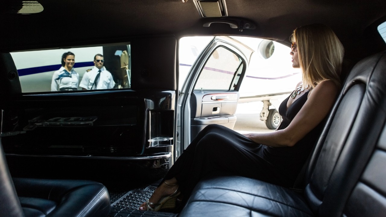 Una donna in abito nero siede in un'auto lussuosa, di fronte a una TV che mostra due persone in divisa da pilota. La portiera dell'auto è aperta, rivelando un jet privato parcheggiato fuori. La scena trasmette eleganza e viaggio.