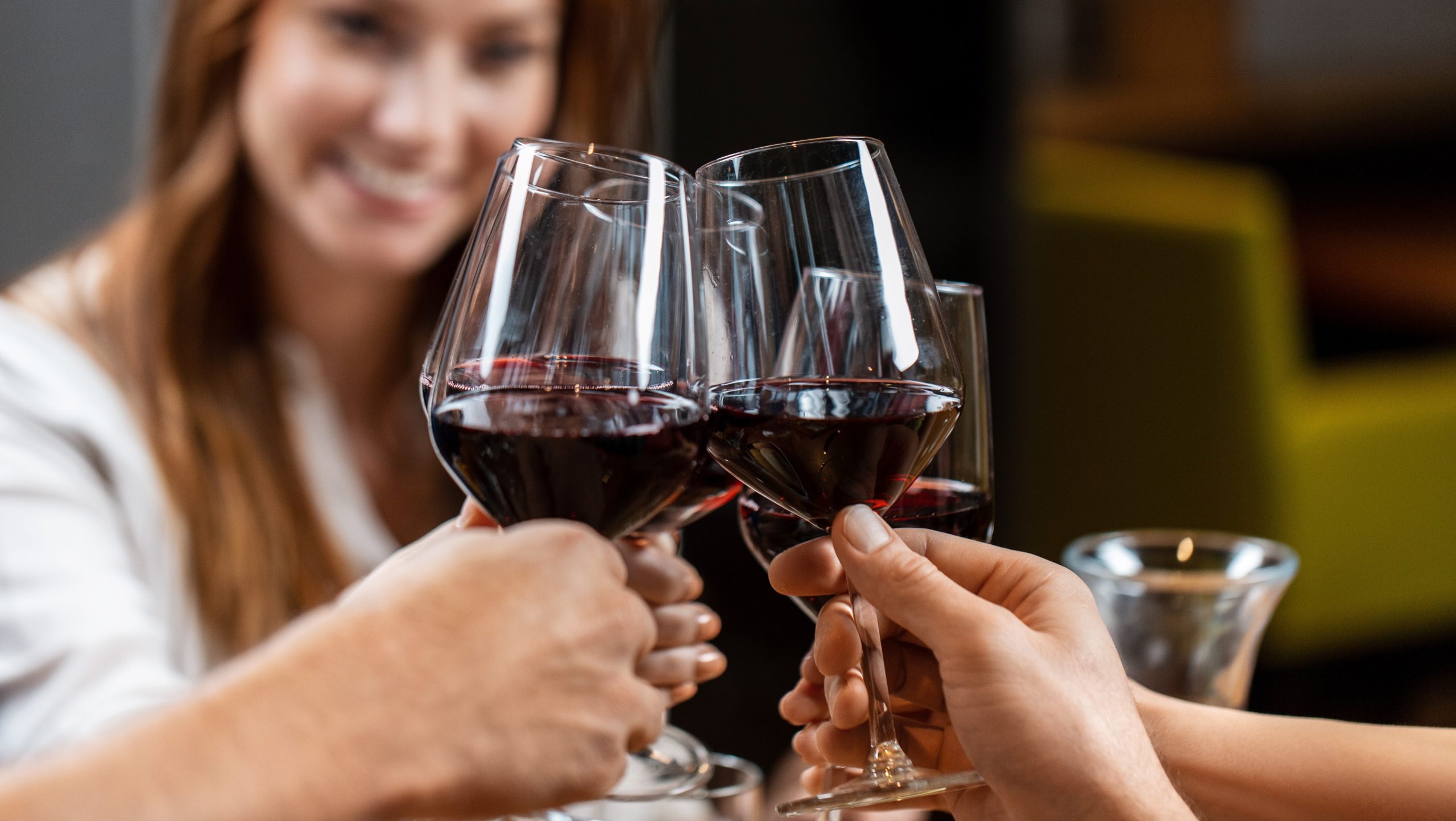 Un gruppo di persone che brindano con bicchieri di vino rosso in un gesto celebrativo. Una donna è visibile mentre sorride sullo sfondo, mentre le mani di altri tengono i bicchieri in primo piano. L'ambiente sembra essere informale e ben illuminato.
