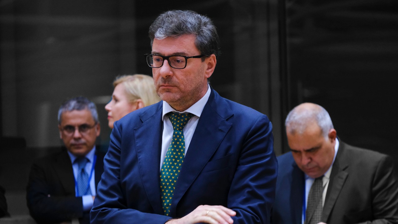 Un uomo in abito blu e cravatta verde a pois è in piedi con un'espressione seria, circondato da altre tre persone in abiti da lavoro, su uno sfondo scuro.