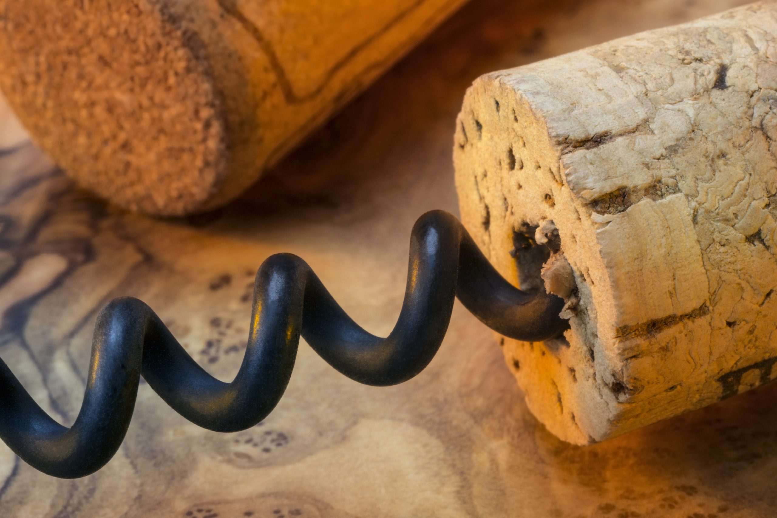 Primo piano di un cavatappi inserito in un tappo di sughero, con un altro tappo sullo sfondo, appoggiato su una superficie di legno. La spirale del cavatappi e la consistenza del tappo sono evidenziate nell'immagine.
